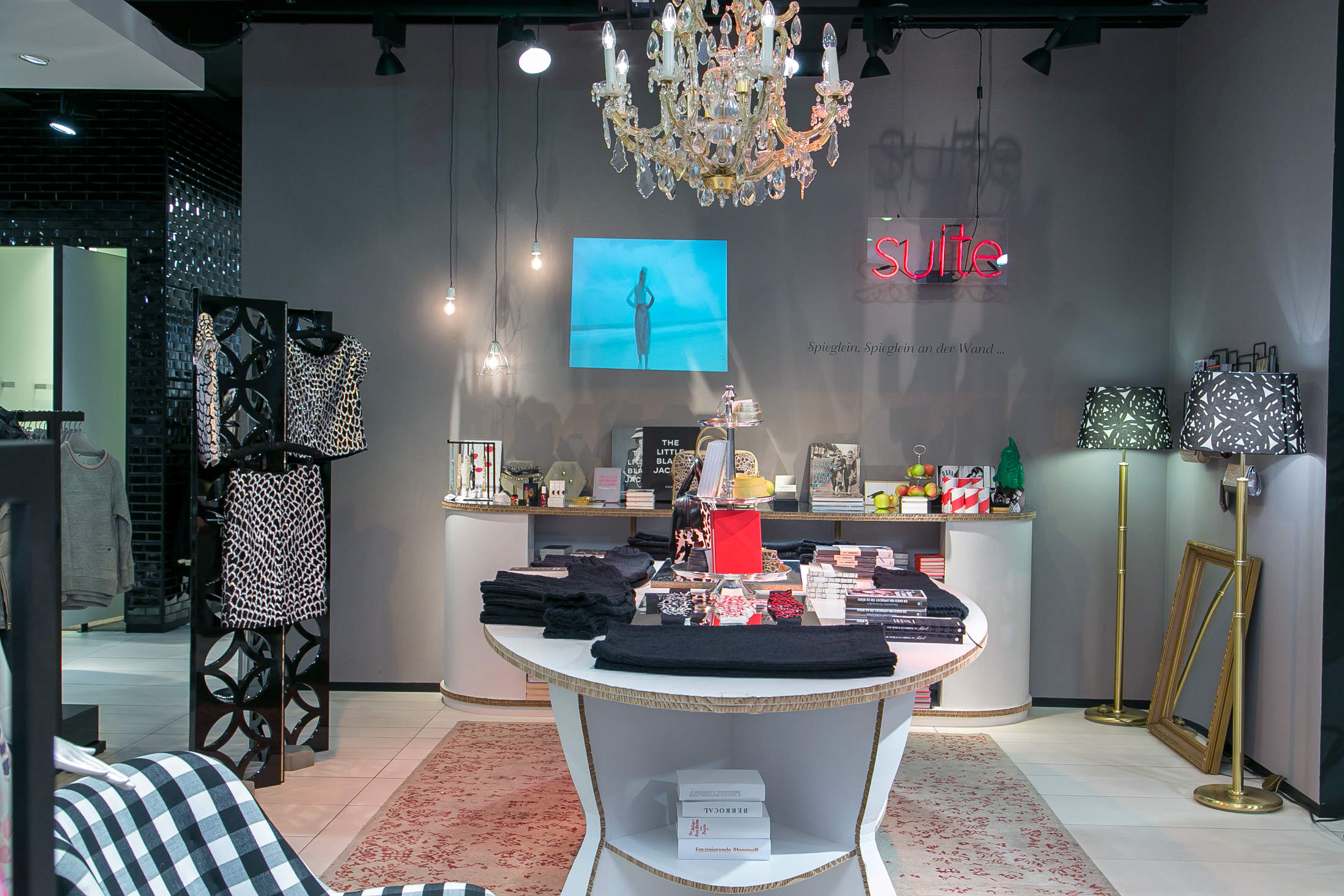 Ein schickes Boutique-Interieur mit einem runden Ausstellungstisch voller gefalteter Kleidung und Accessoires. Darüber hängt ein Kronleuchter und elegante Wanddekorationen, darunter ein gerahmtes Bild und ein Neonschild, schmücken den Raum. Schwarz-weiß gemusterte Gegenstände verleihen Stil.