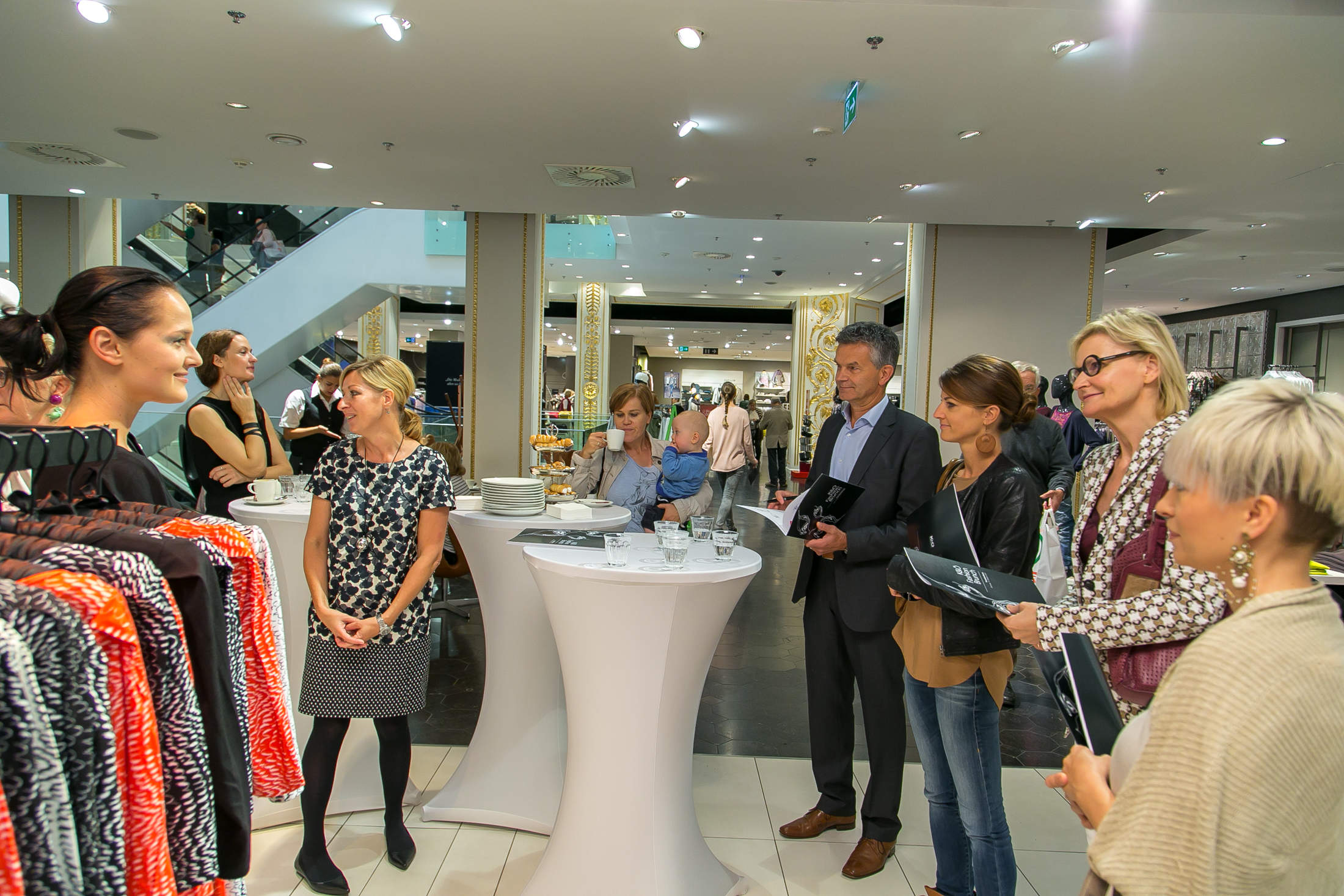 Eine Gruppe von Menschen steht in einem hell erleuchteten Bekleidungsgeschäft und unterhält sich. Mehrere Kleiderständer mit bunter Kleidung sind zu sehen, und auf zwei hohen Tischen stehen Tassen und Teller, was auf ein zwangloses Treffen oder eine Veranstaltung schließen lässt.