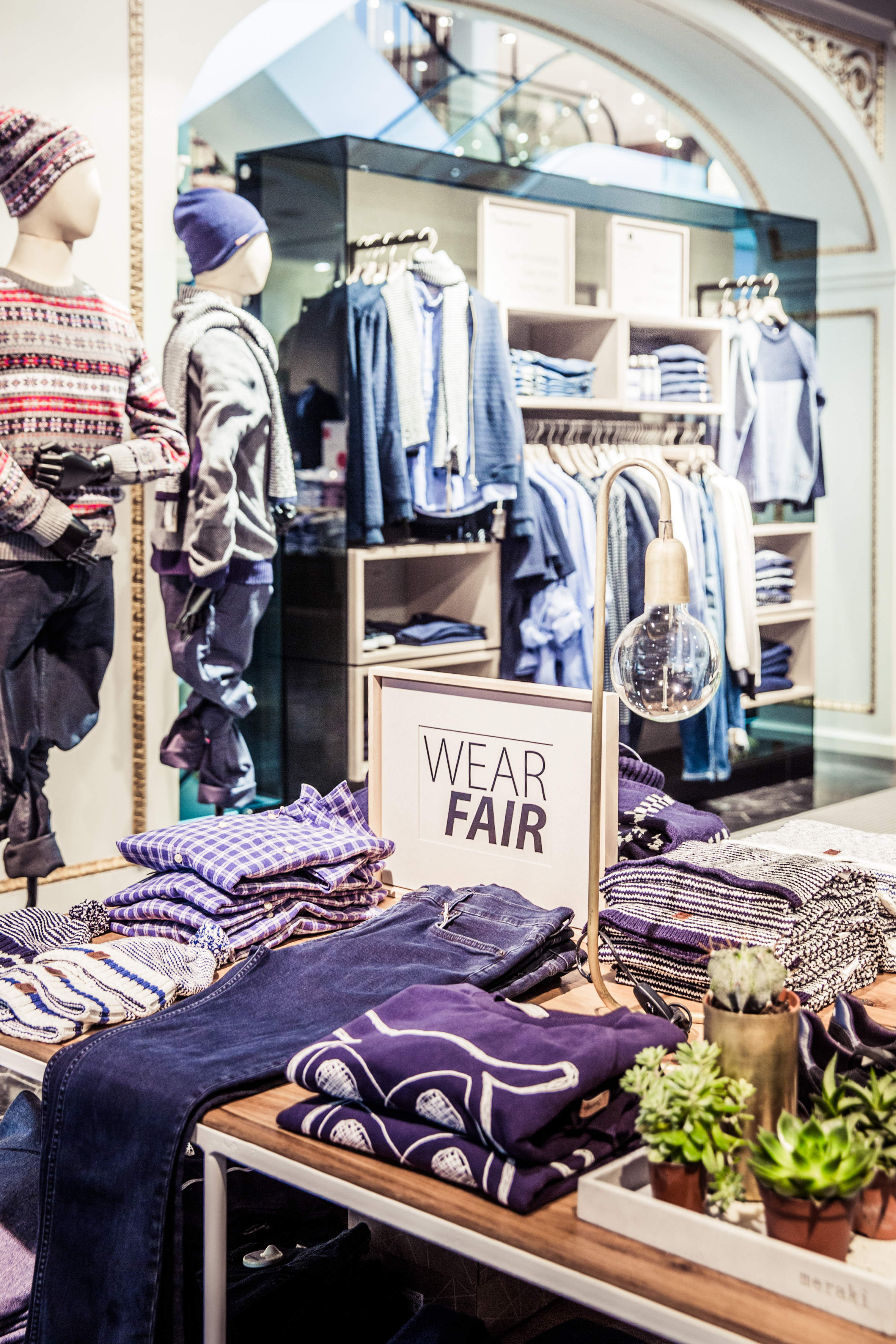 In einem Bekleidungsgeschäft werden gefaltete Hemden und Jeans ausgestellt. Auf einem Schild steht „WEAR FAIR“ und betont Fair-Trade-Mode. In der Nähe stehen Schaufensterpuppen in Freizeitkleidung. Eine hängende Glühbirne und kleine Pflanzen ergänzen das Dekor.