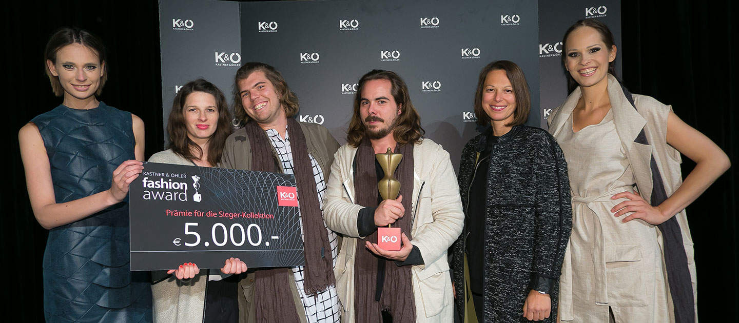 Eine Gruppe von sechs Personen lächelt und hält einen großen Scheck und eine Trophäe in den Händen. Sie stehen vor einem Hintergrund mit den Worten „K&O“ und „Fashion Award“. Auf dem Scheck steht „5.000 €“ als Preissumme.