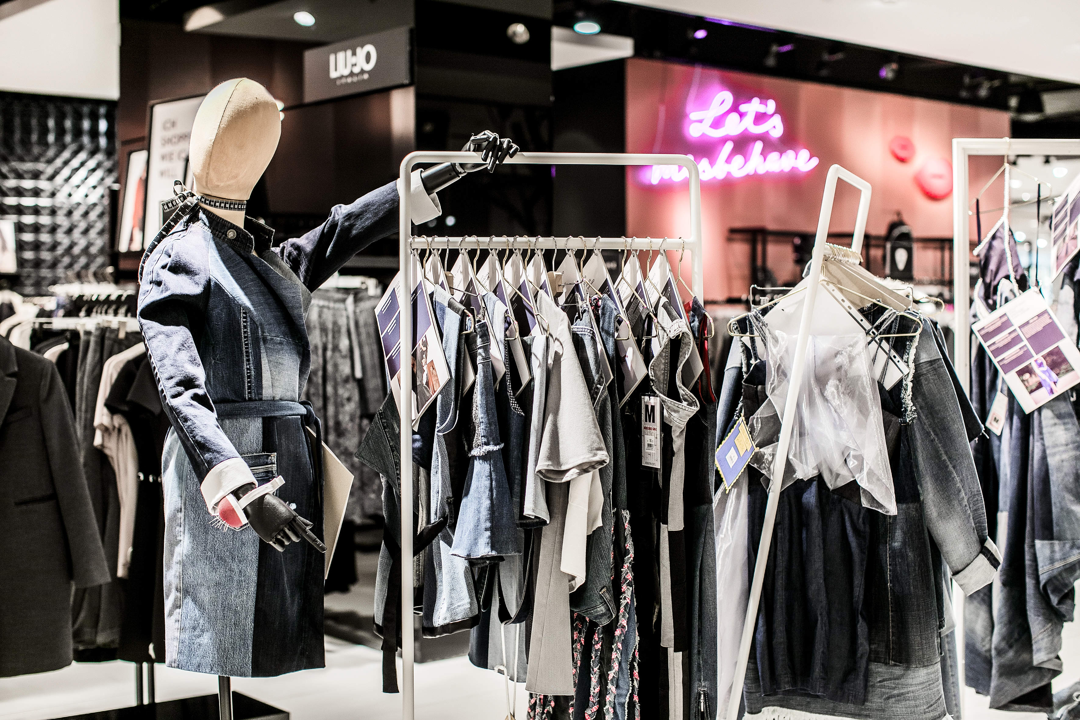 Ein Schaufenster in einem Einzelhandelsgeschäft mit einer Schaufensterpuppe, die eine Jeansjacke trägt. Mehrere Regale präsentieren Jeans-Outfits und -Kleidung. Im Hintergrund steht auf einem Neonschild „Lasst uns netzwerken“. Das Ambiente ist stilvoll und modern.