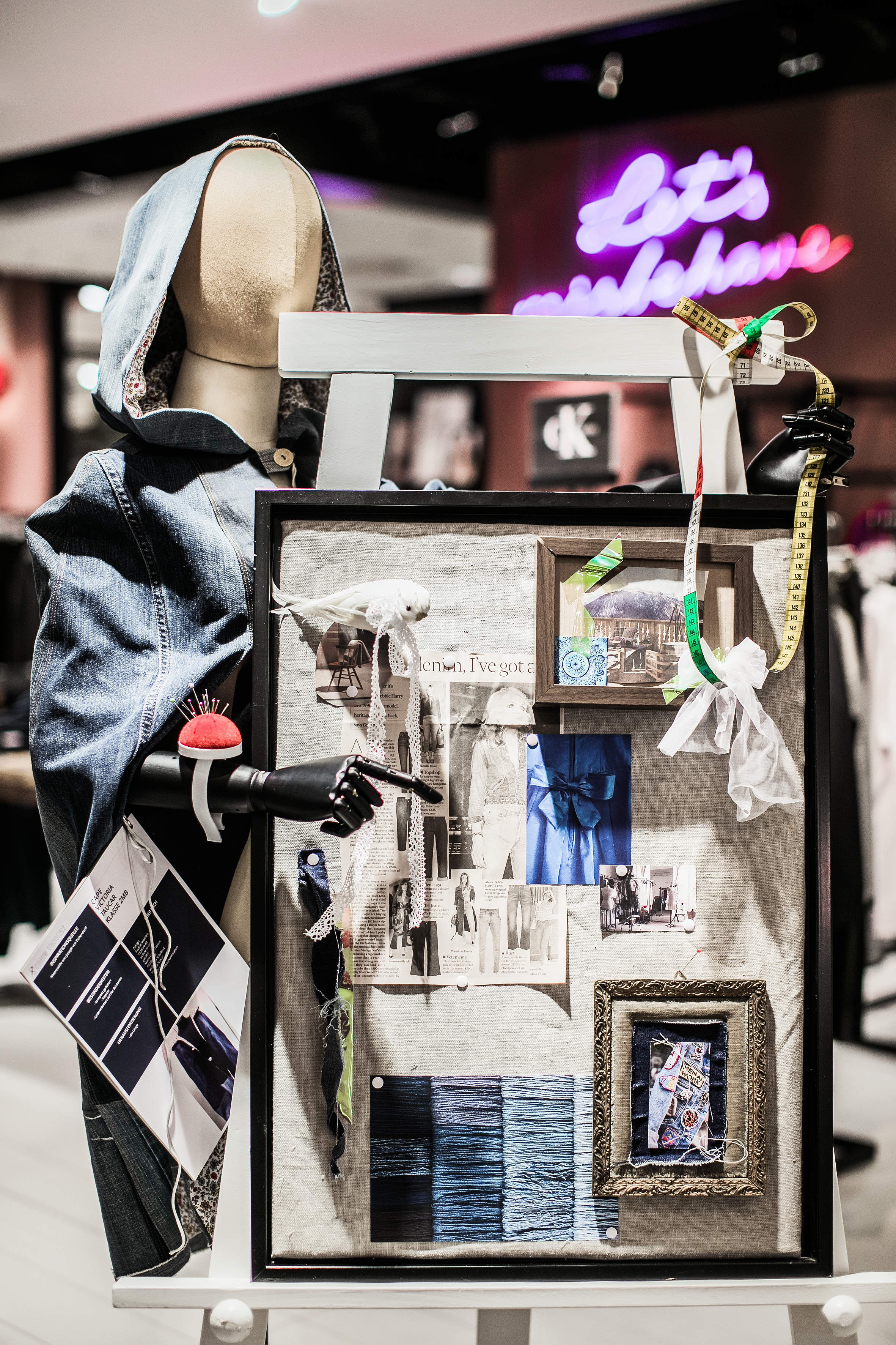 Eine Schaufensterpuppe im Jeans-Outfit steht neben einer künstlerischen Tafel, die mit Modeskizzen, Stoffmustern und gerahmten Fotos geschmückt ist. An der Tafel sind ein Maßband und eine Schere befestigt. Im Hintergrund ist ein Neonschild zu sehen.