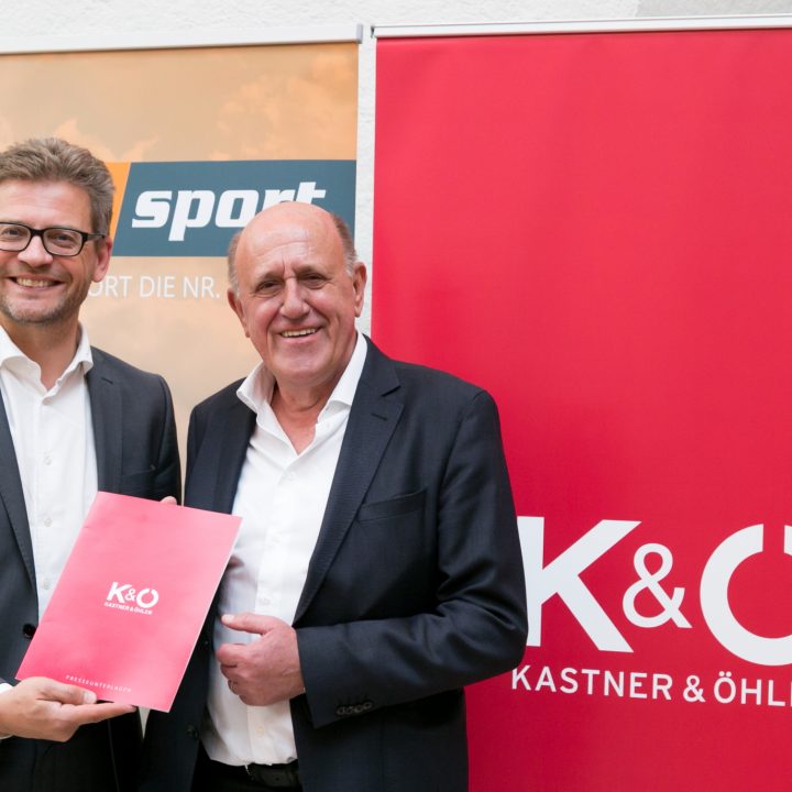 Zwei Männer in Anzügen stehen lächelnd da. Einer hält einen roten Ordner mit der Aufschrift „K&O Kastner & Öhler“. Dahinter hängen Banner mit den Logos von Sky Sport und Kastner & Öhler.