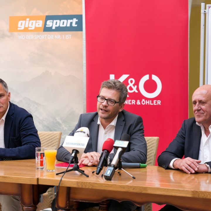 Drei Männer in Anzügen sitzen an einem Konferenztisch mit Mikrofonen und geben eine Pressekonferenz. Dahinter hängen Banner von „giga sport“ und „K&Ö KARNER & ÖHLER“. Rechts ist eine bunte Grafik teilweise zu sehen.