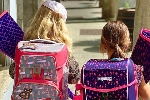 Zwei Kinder gehen auf einem sonnigen Weg und tragen jeweils bunte Rucksäcke und Ordner. Das linke Kind hat einen rosa Rucksack mit Baummustern und das rechte Kind einen dunklen Rucksack mit violetten Mustern. Sie gehen in Richtung eines schattigen Bereichs.