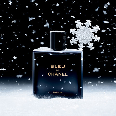 Eine Flasche Bleu de Chanel-Parfüm steht aufrecht auf einer schneebedeckten Oberfläche. Schneeflocken fallen sanft um sie herum und eine große, dekorative Schneeflocke lehnt an der Flasche. Der Hintergrund ist dunkel und hebt das Produkt hervor.