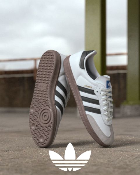 Ein Paar Adidas Samba-Sneaker mit weißem Obermaterial, schwarzen Streifen und Gummisohle wird im Freien ausgestellt. Ein Schuh steht aufrecht, der andere ist geneigt, sodass die Sohle zu sehen ist. Unten ist das Adidas-Logo zu sehen.