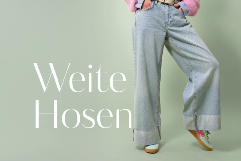 Person in Jeans mit weitem Bein und rosa Pullover posiert vor hellgrünem Hintergrund. Links ist der Schriftzug „Weite Hosen“ zu sehen. Grüne und rosa Turnschuhe sind zu sehen.