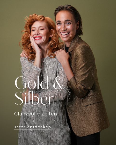 Zwei lächelnde Menschen stehen dicht beieinander vor einem grünen Hintergrund. Die eine trägt ein funkelndes silbernes Kleid, die andere einen glänzenden goldenen Blazer. Der Text lautet: „Gold & Silber Glanzvolle Zeiten jetzt entdecken.“