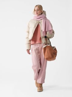 Eine Person in rosa und beiger Winterkleidung steht vor einem weißen Hintergrund. Sie trägt eine Pufferjacke, einen Schal, einen Pullover und weite Hosen mit Stiefeln und trägt eine braune Tasche. Der Gesamteindruck ist gemütlich und modisch.