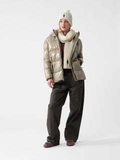 Eine Person, die eine glänzende, beige Pufferjacke, eine cremefarbene Beanie-Mütze, einen passenden Schal, schwarze Hosen mit weitem Bein und braune Schuhe trägt, steht vor einem schlichten weißen Hintergrund.