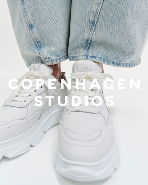 Nahaufnahme von weißen Turnschuhen mit dicken Sohlen, die von einer Person in hellblauen Jeans getragen werden. Der Text „COPENHAGEN STUDIOS“ ist über das Bild gelegt.