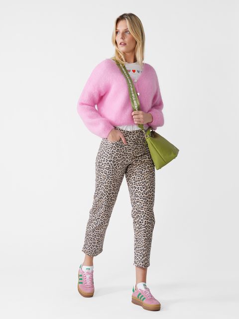 Eine Frau in einem rosa Pullover, einer Hose mit Leopardenmuster und rosa Turnschuhen steht vor einem weißen Hintergrund. Sie trägt eine grüne Umhängetasche und blickt zur Seite.