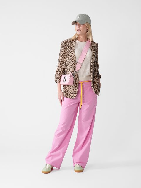 Eine Person trägt einen Blazer mit Leopardenmuster, einen beigen Pullover und eine leuchtend rosa Hose. Sie trägt eine hellgraue Mütze und eine rosa Umhängetasche. Sie steht vor einem schlichten weißen Hintergrund und blickt zur Seite.