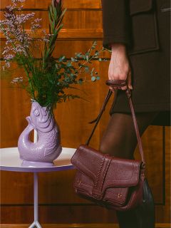 Eine Person in dunkler Kleidung hält eine burgunderrote Handtasche neben einer lila, fischförmigen Vase mit Blumen auf einem weißen Tisch.