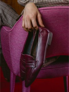 Eine Person in karierten Hosen sitzt auf einem rosa Stuhl mit strukturierter Oberfläche und hält einen burgunderroten Slipper in der Hand.