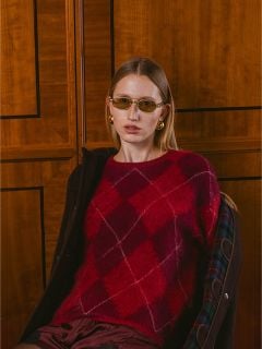 Eine Frau, die einen roten Argyle-Pullover, eine Sonnenbrille und goldene Ohrringe trägt, sitzt vor einer Wand mit Holzvertäfelung und blickt in die Kamera.