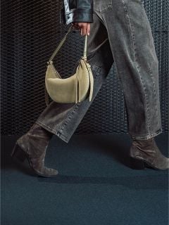 Eine Person in grauen Jeans und Wildlederstiefeln mit Absatz hält eine beige Handtasche in der Hand und geht vor einem strukturierten dunklen Hintergrund vorbei.