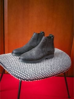 Ein Paar schwarze Chelsea-Stiefel auf einem schwarz-weiß gemusterten Stuhl vor einem roten Teppich und einer Holzwand.