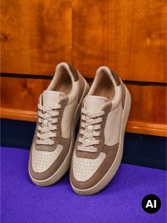 Ein Paar beige-braune Sneaker mit weißen Schnürsenkeln steht vor einem hölzernen Hintergrund auf einem violetten Boden.