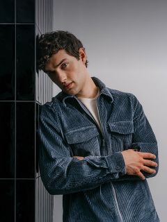Ein Mann, der ein blaues Cordhemd mit Reißverschluss trägt, die Arme verschränkt hat und an einer dunkel gefliesten Wand lehnt.